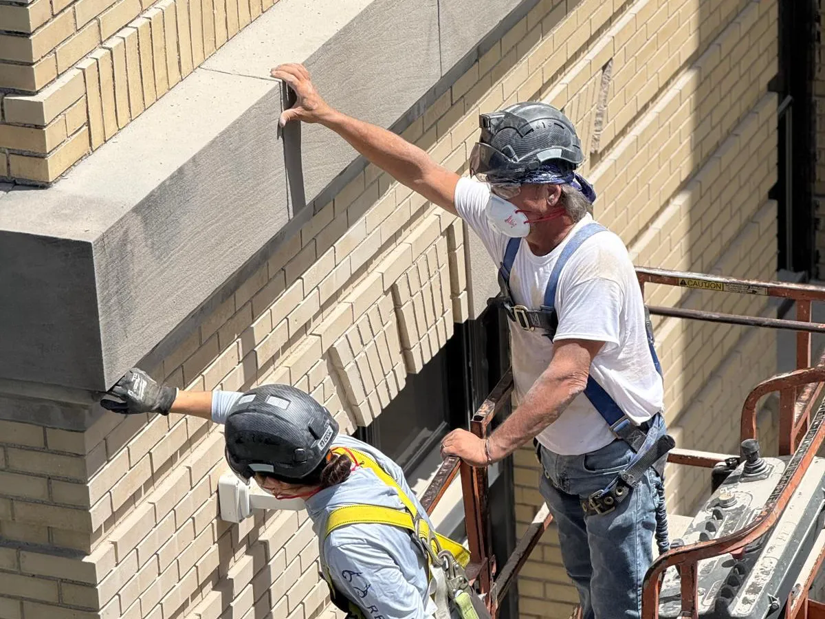 Two Bramwell-McKay technicians in helmets and harnesses on a lift, working along the stone band of a cream brick building